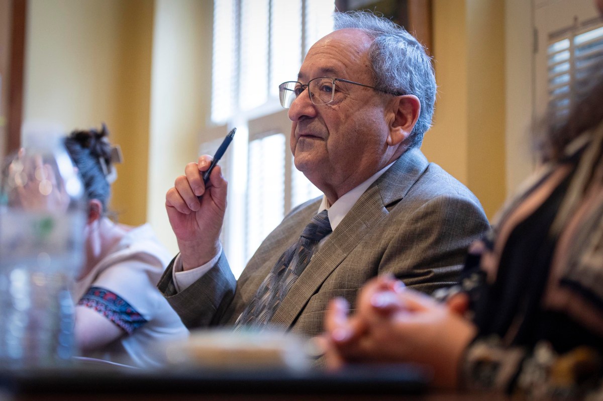 An older man in a suit holds a pen and listens attentively during an indoor meeting, seated at a table with other people.