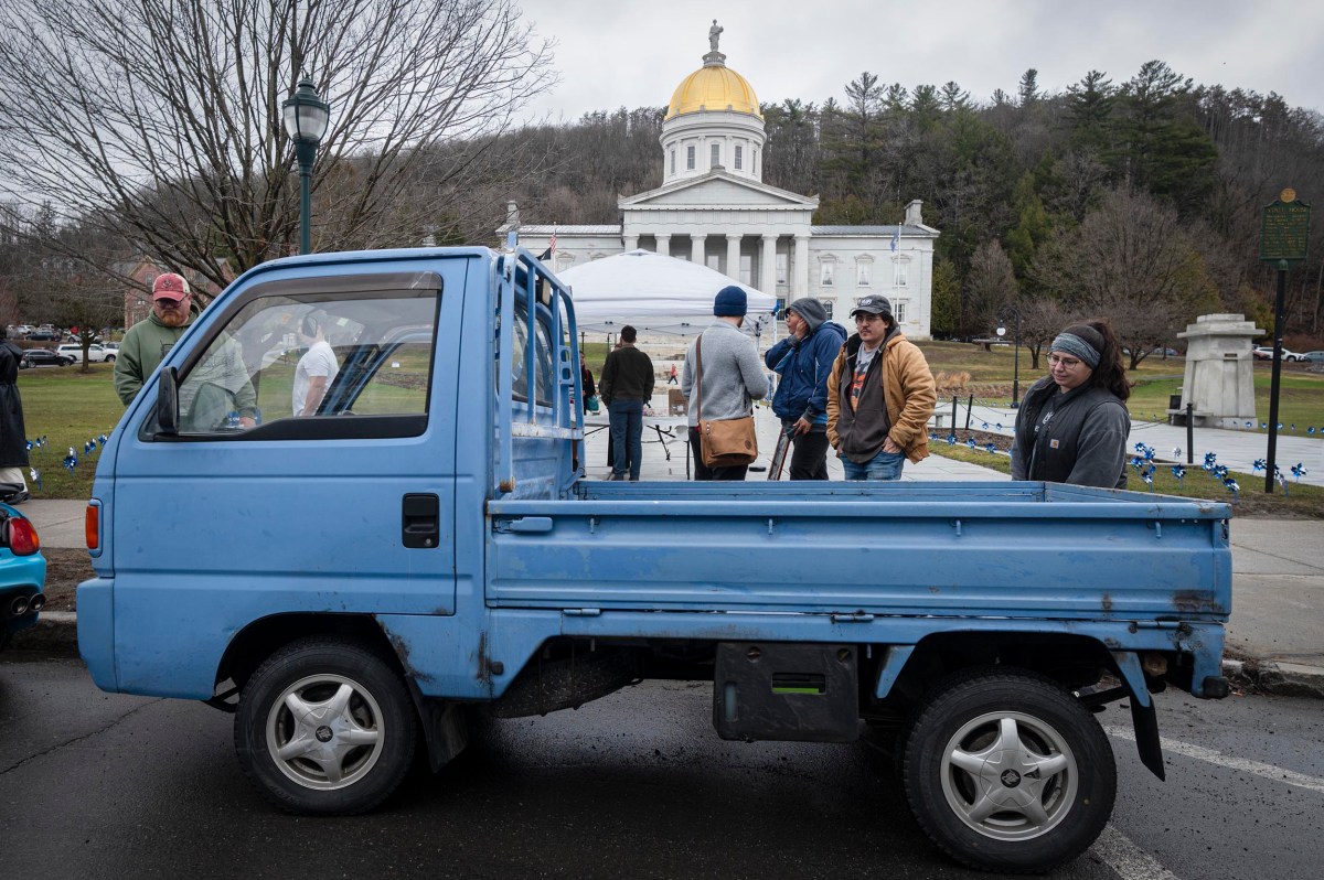 A group of people stand near a small blue pickup truck parked in front of a domed capitol building on a cloudy day.
