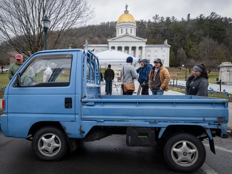 A group of people stand near a small blue pickup truck parked in front of a domed capitol building on a cloudy day.