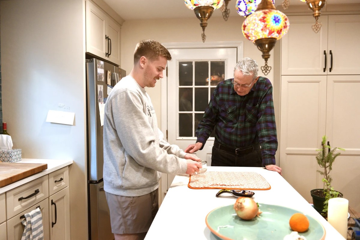 Two people stand in a kitchen; one is preparing food on a cutting board while the other observes. Colorful pendant lights hang above the island.