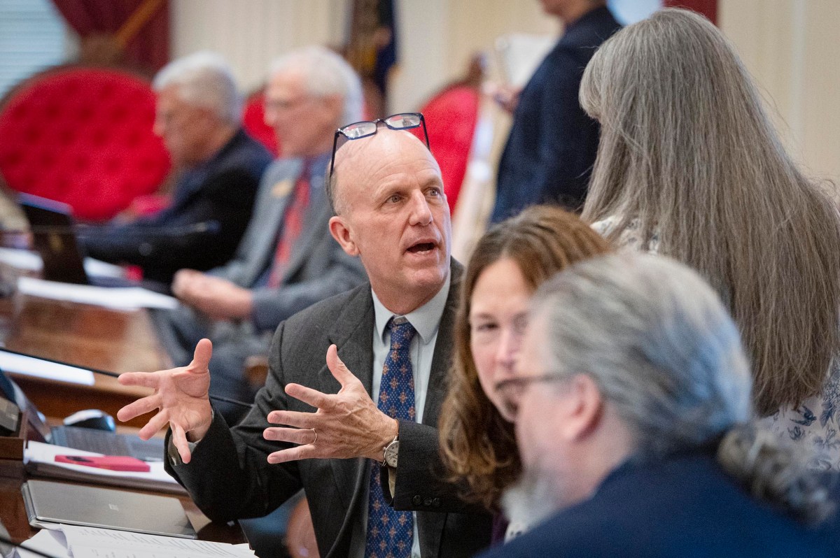 A man in a suit talks animatedly with colleagues in a formal meeting room, gesturing with his hands; others listen and work at desks in the background.