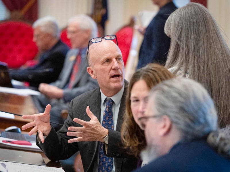 A man in a suit talks animatedly with colleagues in a formal meeting room, gesturing with his hands; others listen and work at desks in the background.