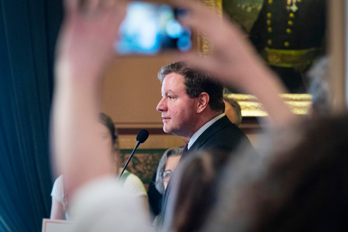 A man in a suit speaks into a microphone at an indoor event, while someone in the foreground holds up a phone to take a photo or video.