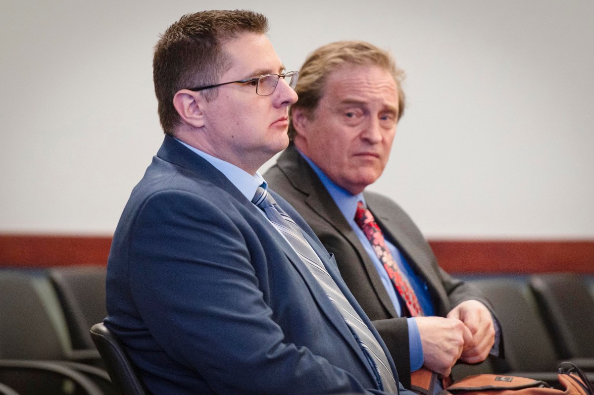 Two men in suits sit next to each other in a row of chairs, one looking forward and the other glancing to the side, in what appears to be a formal setting.