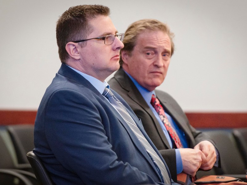 Two men in suits sit next to each other in a row of chairs, one looking forward and the other glancing to the side, in what appears to be a formal setting.