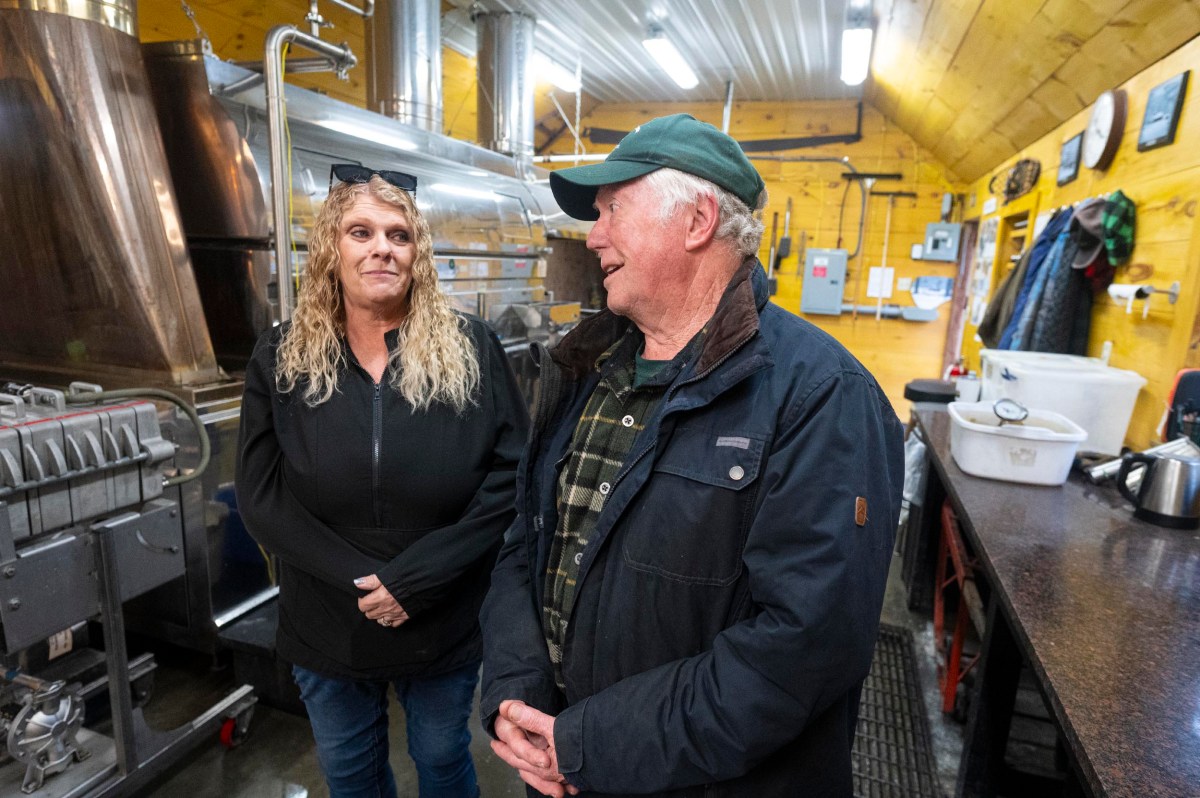 Two people stand and talk inside an industrial kitchen or workshop with metal equipment and yellow wooden walls.