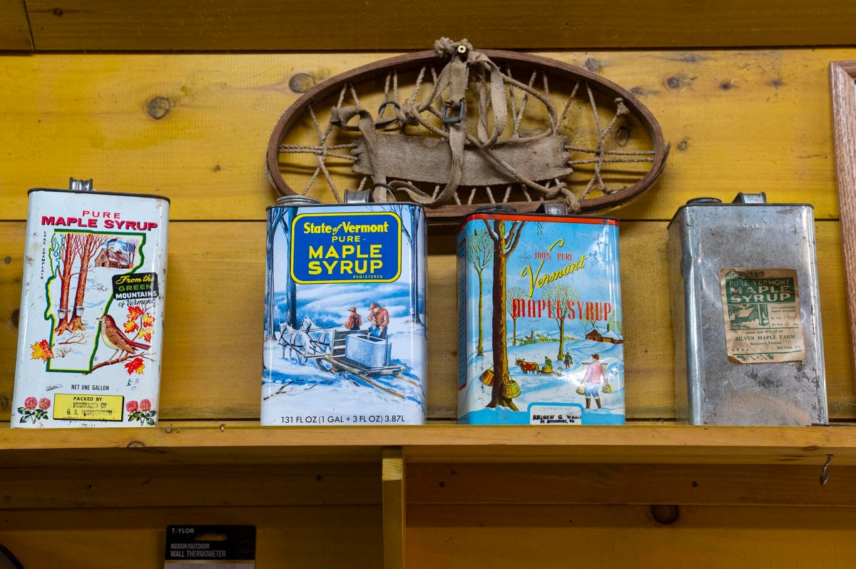 Four vintage maple syrup cans with colorful labels are displayed on a wooden shelf beneath a small antique snowshoe mounted on a yellow wall.