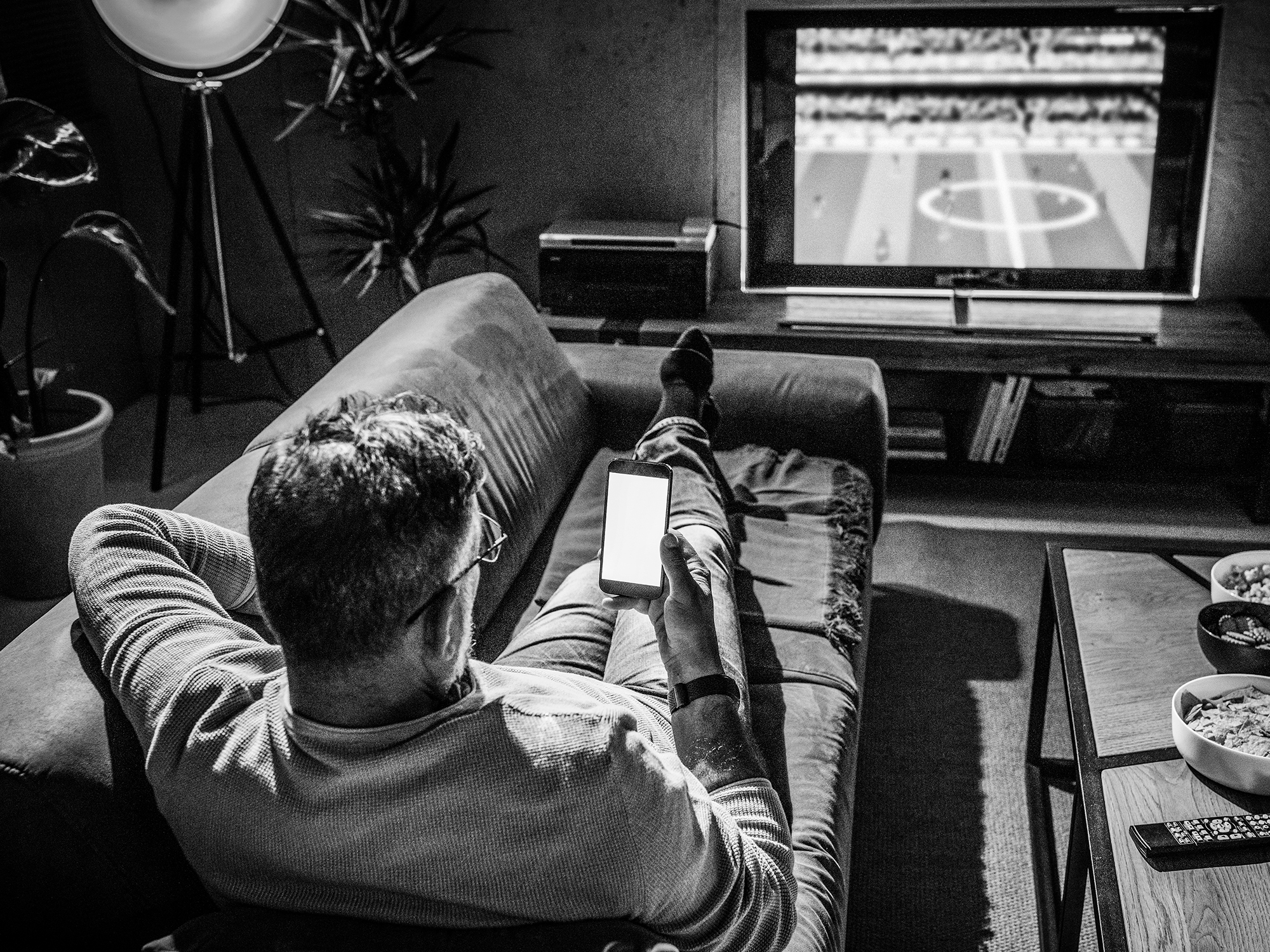 A person sits on a sofa, looking at a smartphone while watching a soccer match on TV; snacks and a remote are on the table nearby.