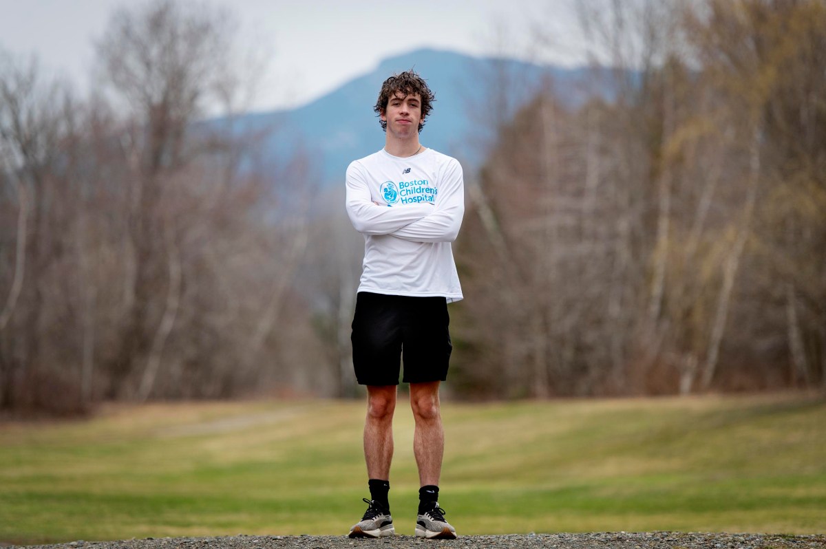 A person stands outdoors on a gravel path with arms crossed, wearing a white long-sleeve shirt, black shorts, and sneakers. Trees and mountains are visible in the background.