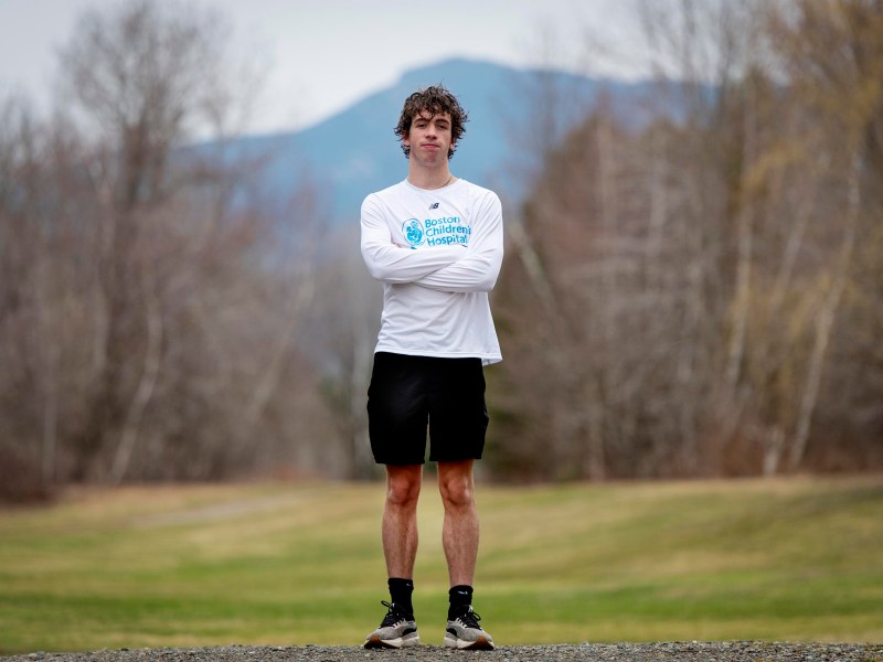 A person stands outdoors on a gravel path with arms crossed, wearing a white long-sleeve shirt, black shorts, and sneakers. Trees and mountains are visible in the background.