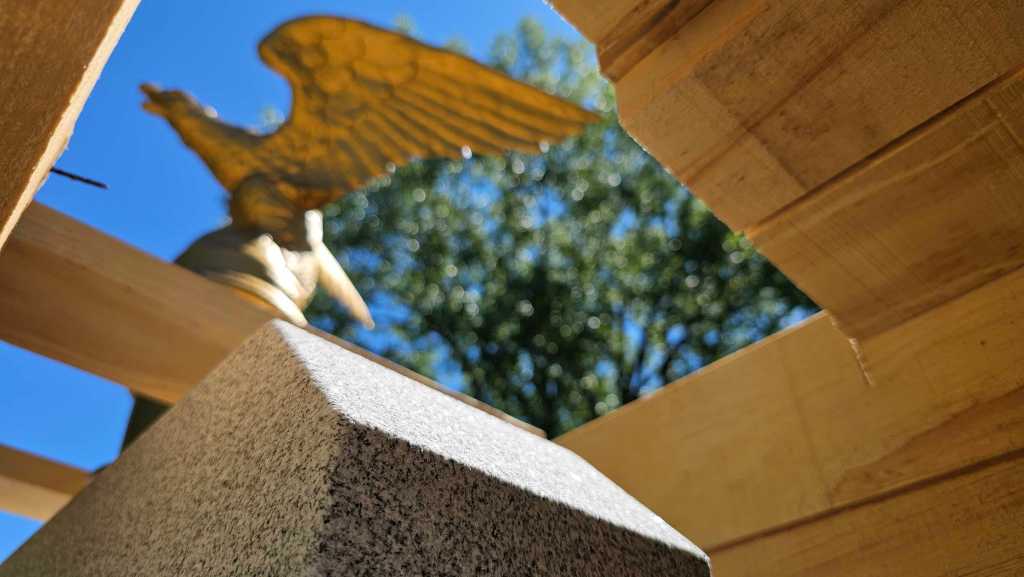 Close-up of a stone block framed by wooden beams, with a gold eagle statue and trees visible in the blurred background under a clear blue sky.