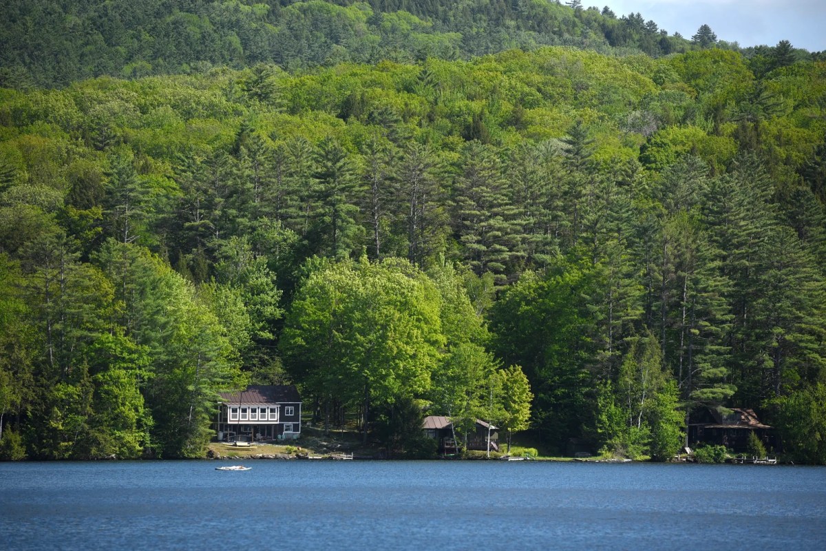 A house and small docks are nestled among dense green trees on the shore of a calm lake, with forested hills in the background.