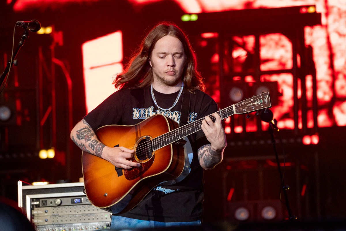 A musician with long hair and tattoos plays an acoustic guitar on stage, with stage equipment and red lighting in the background.