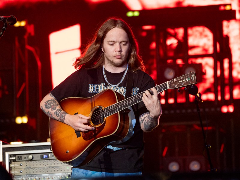 A musician with long hair and tattoos plays an acoustic guitar on stage, with stage equipment and red lighting in the background.