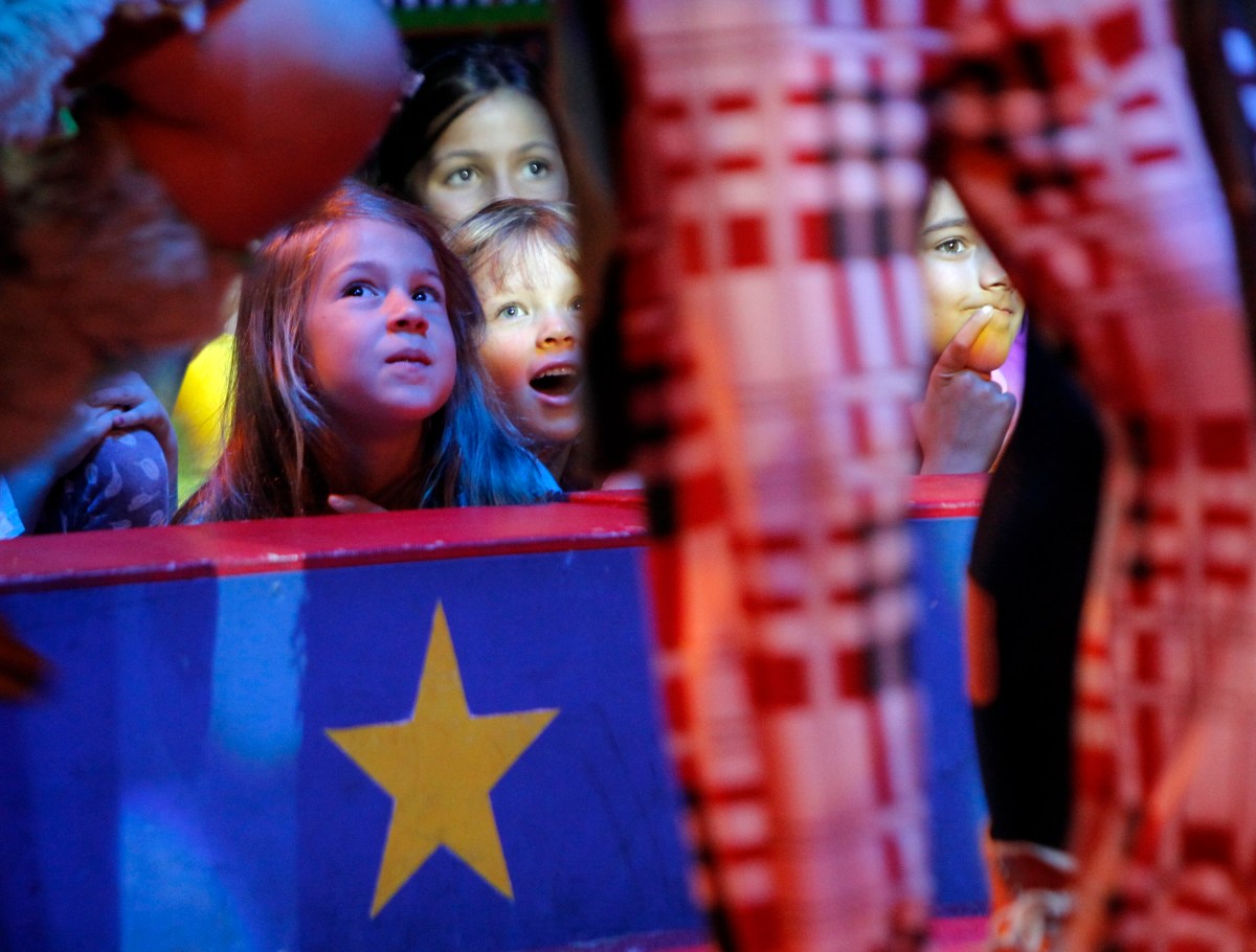 Several children sit behind a barrier decorated with a yellow star, looking up in awe at a performer in plaid pants standing in front of them.