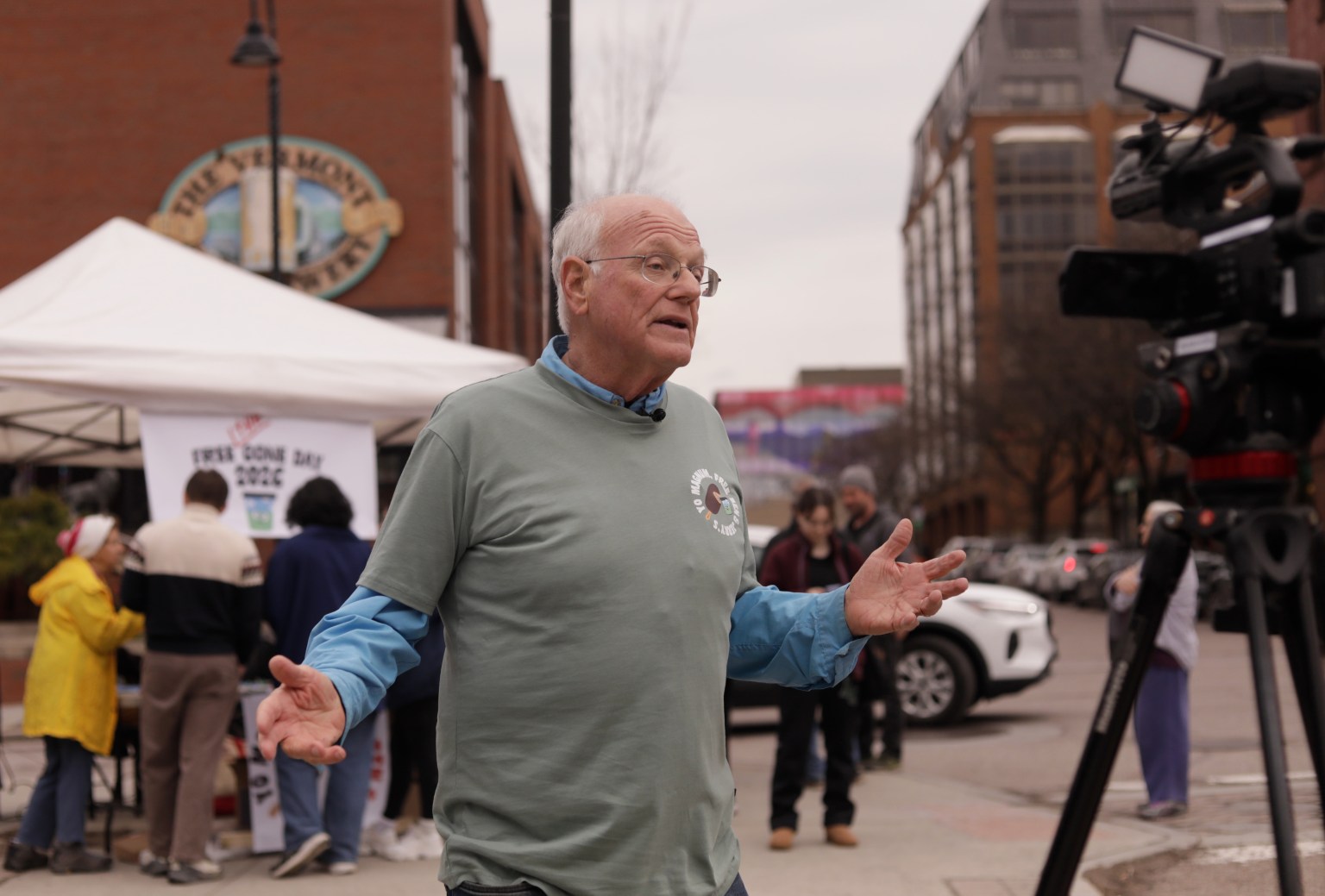 An older man speaks in front of a camera on a city sidewalk, with people gathered near a white tent and brick buildings in the background.