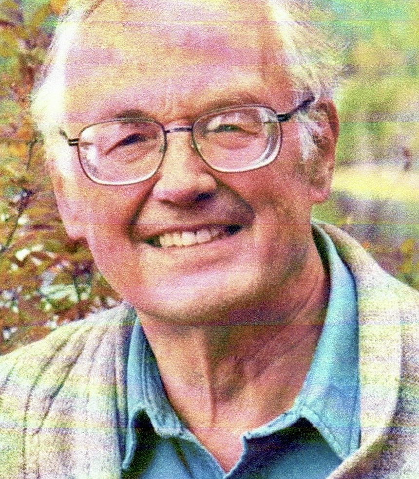 An older man with glasses and a plaid jacket smiles outdoors in natural light, with plants in the background.