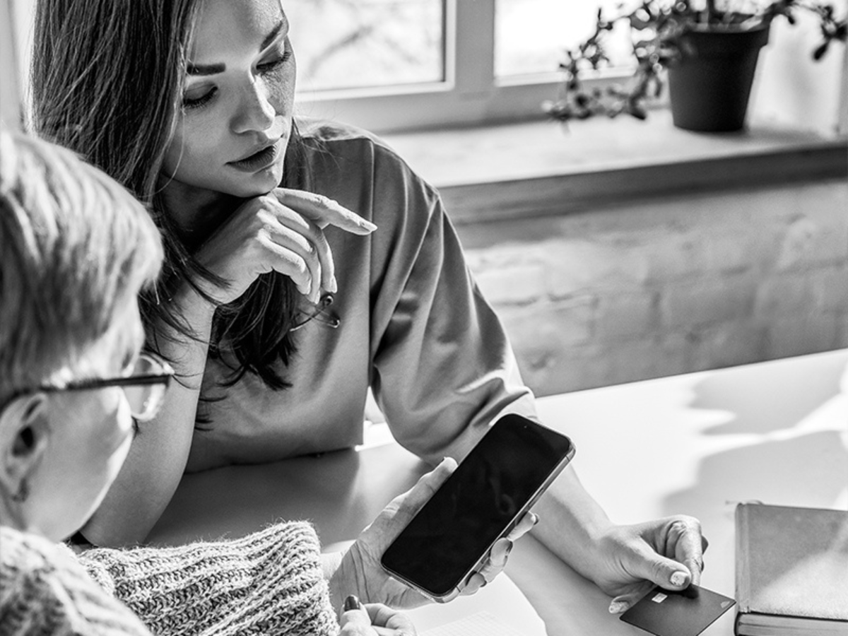Two women sit at a table; one holds a smartphone and credit card while the other points at the phone screen.