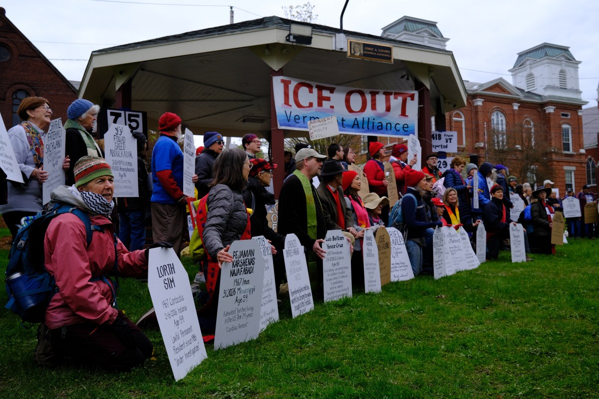 A group of people gathered outside holding signs and banners that read "ICE OUT Vermont Alliance," protesting near a gazebo and historic brick buildings.