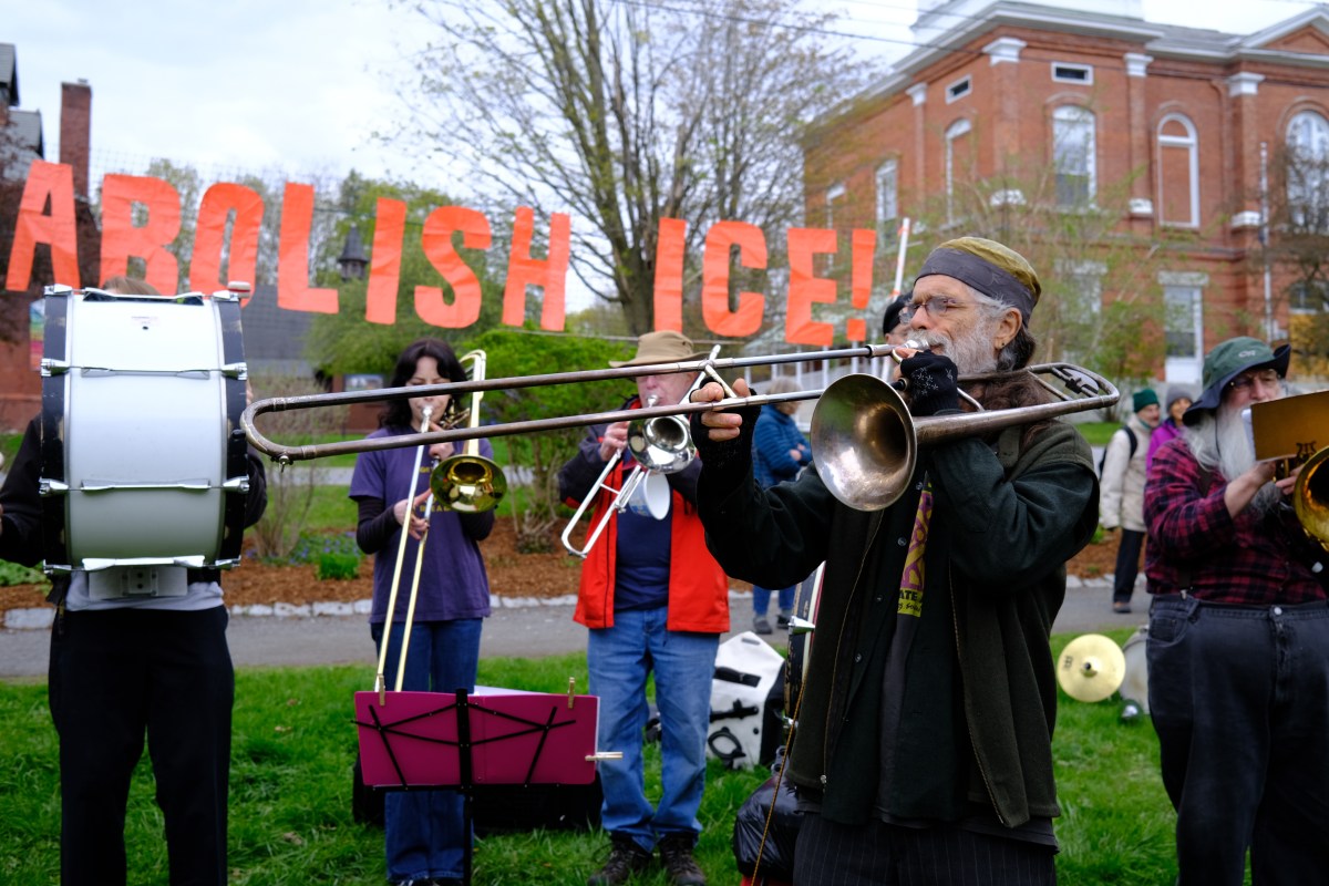 A group of musicians play instruments outdoors in front of large orange letters spelling "ABOLISH ICE!.