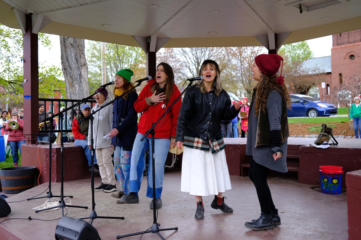 Four women stand on a gazebo stage singing into microphones, wearing casual, colorful clothing and jackets; people watch from the background in an outdoor setting.