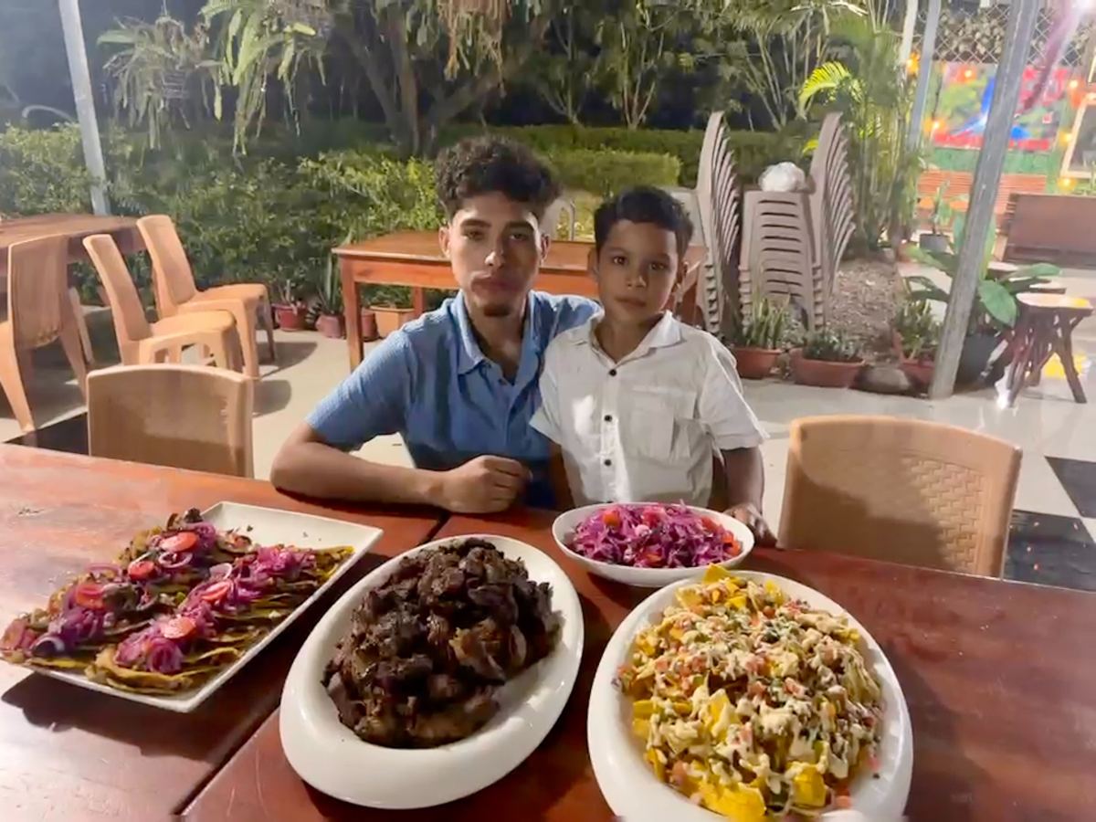 Two boys sit at an outdoor table with three large platters of food and a bowl of salad in front of them. The background features plants, chairs, and tables.