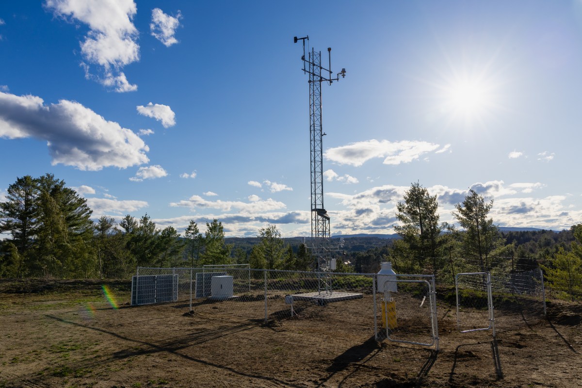 A fenced weather station with a tall metal tower and meteorological instruments stands on a hilltop under a sunny, partly cloudy sky. Trees and distant hills are visible in the background.