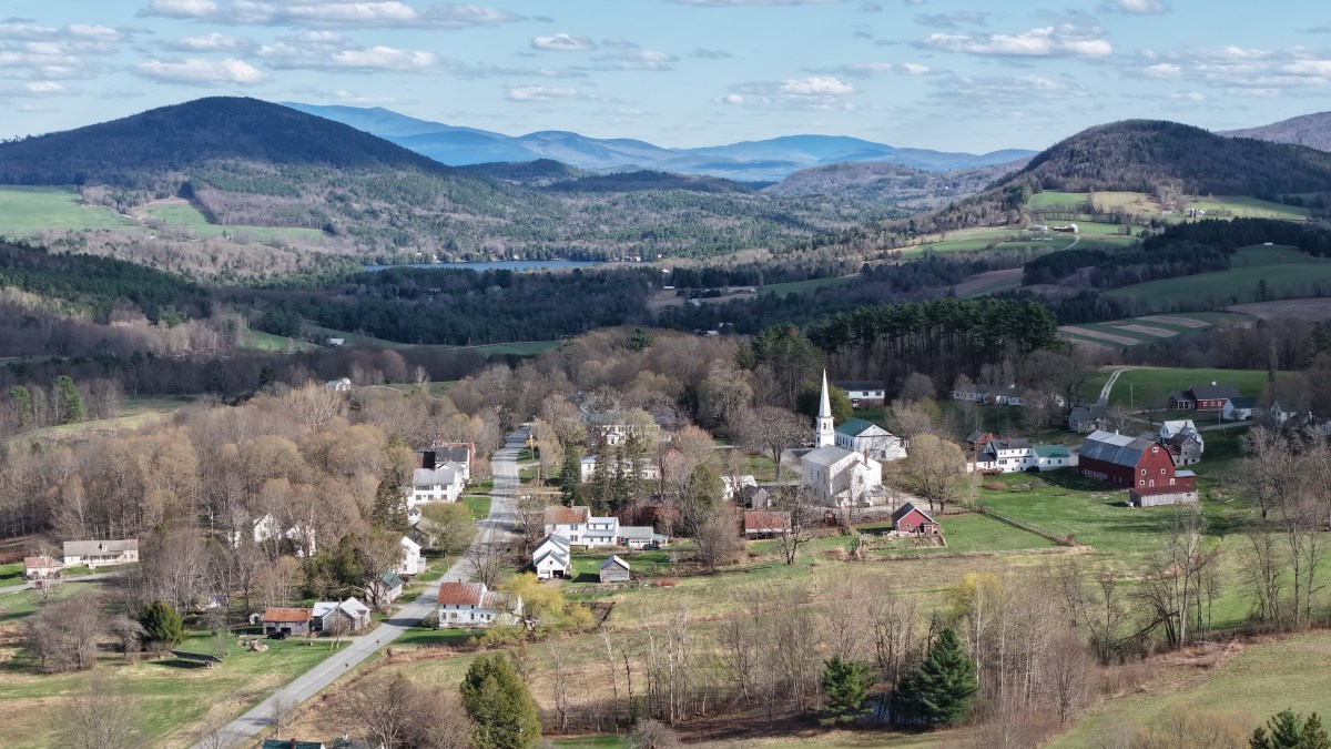 A small rural village with white houses, a church with a steeple, and red barns, surrounded by rolling hills and mountains under a partly cloudy sky.