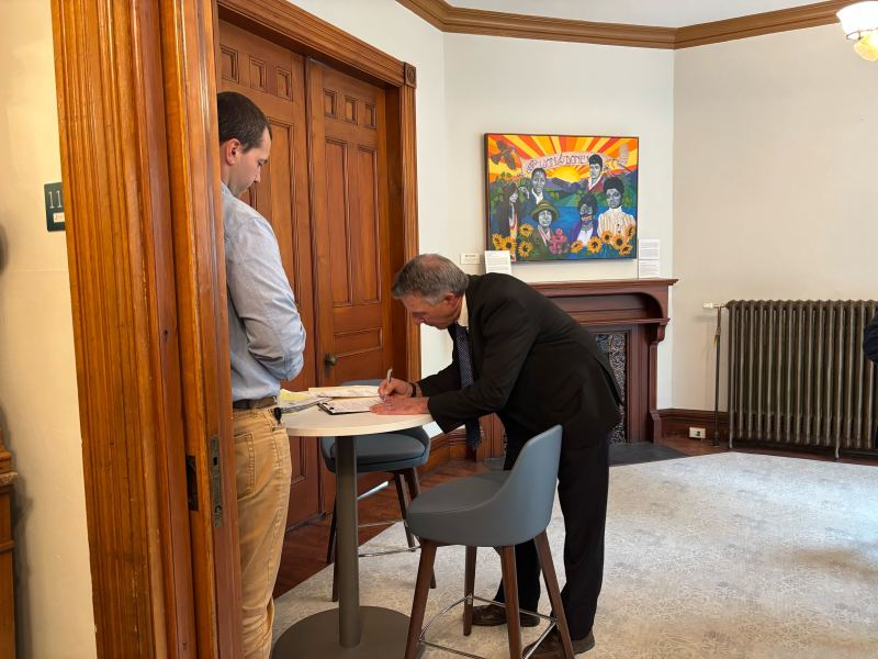 A man in a suit signs a document at a tall table while another man stands nearby in an office with wooden doors and a colorful painting on the wall.