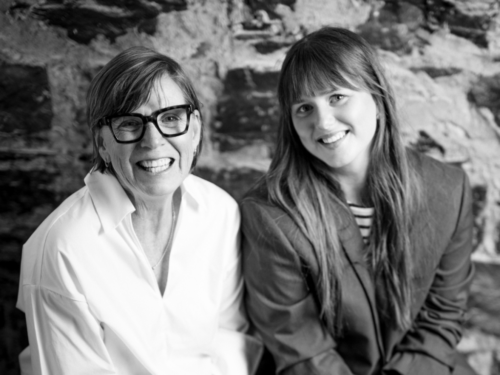 Two women sit side by side, smiling at the camera in front of a stone wall. One wears glasses and a light shirt; the other has long hair and a striped top with a blazer. Black-and-white photo.