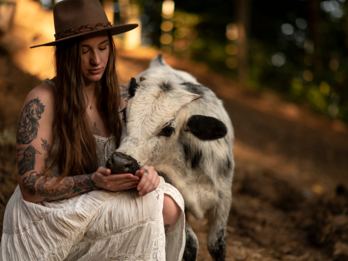 A woman in a white dress and brown hat sits outdoors, feeding a black and white calf from her hand.