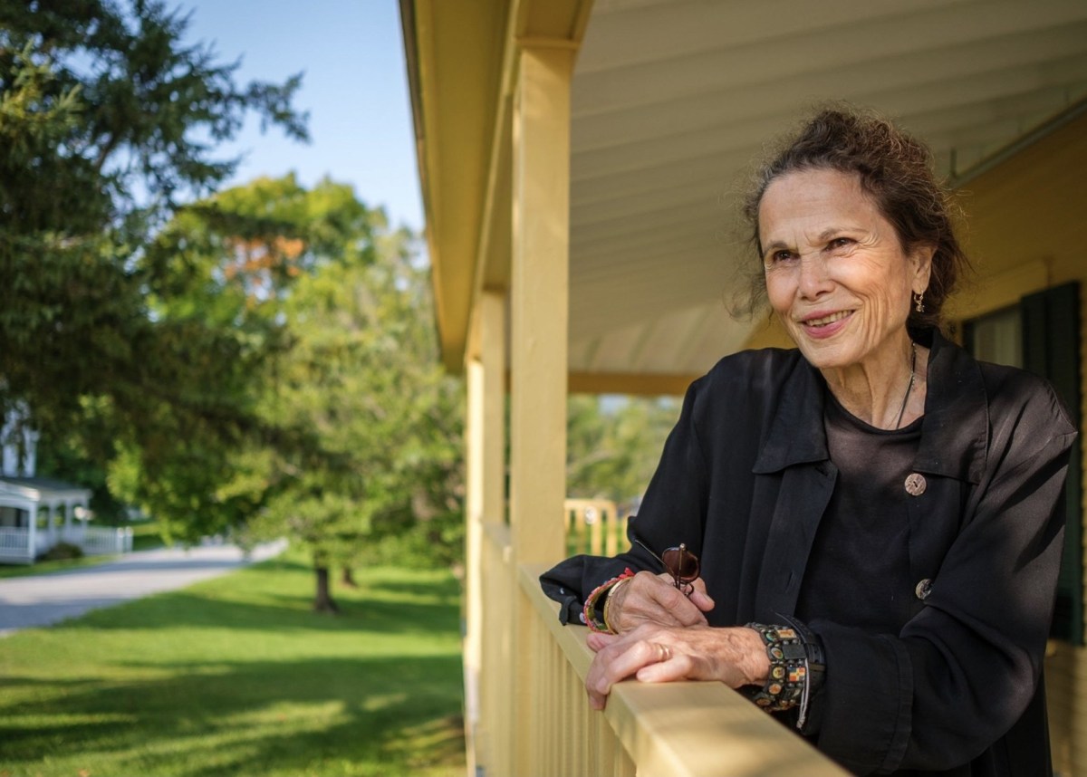 An older woman in a black jacket leans on a yellow porch railing, smiling, with greenery and a house visible in the background.
