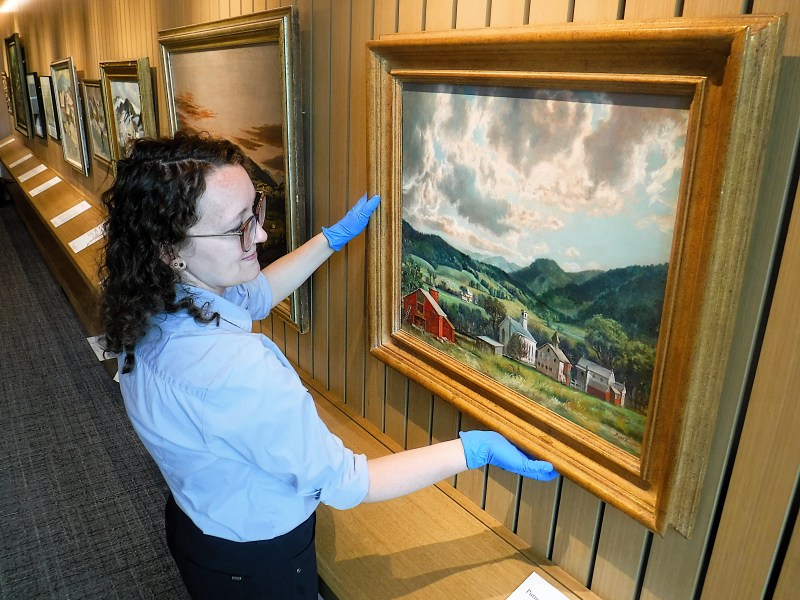 A person wearing blue gloves adjusts a framed landscape painting on a wall in a gallery with several other artworks displayed.