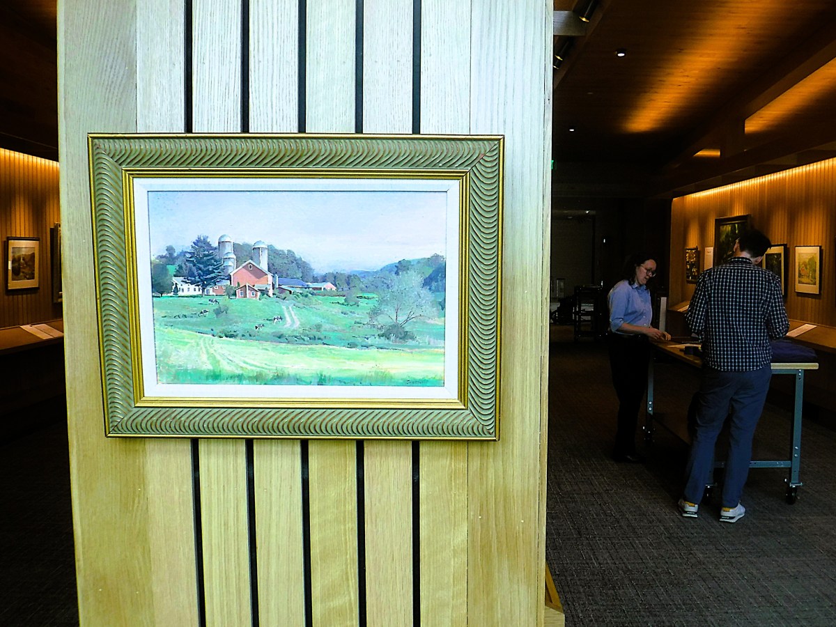 A framed landscape painting of a farm scene hangs on a wooden wall panel; two people stand at a table in the background of a warmly lit room.