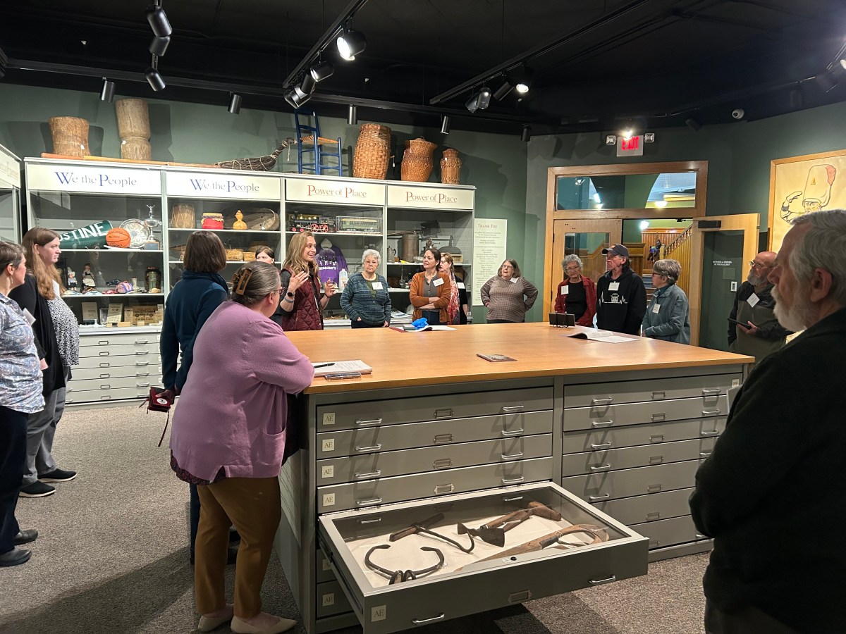 A group of people stands around a large table in a museum or educational setting, listening to a speaker. Cabinets and artifacts are visible in the background.