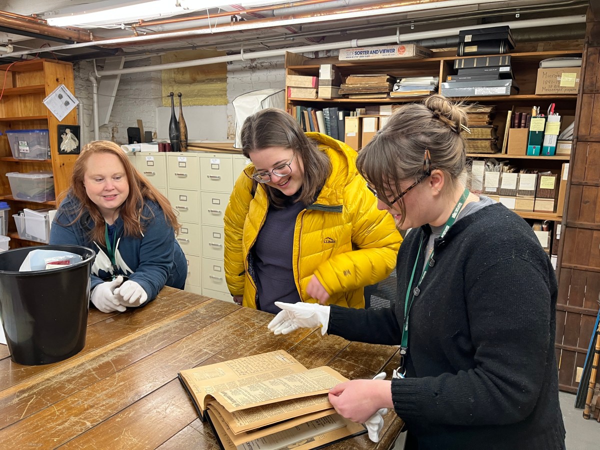 Three women, two wearing gloves, examine an old book at a wooden table in a room filled with shelves, cabinets, and archival materials.