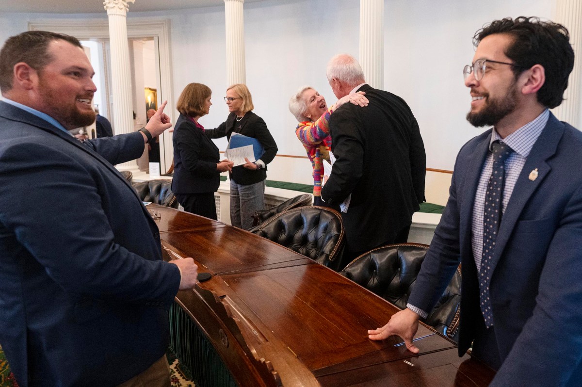 People in business attire interact and converse around a wooden table in a formal room with columns and leather chairs.