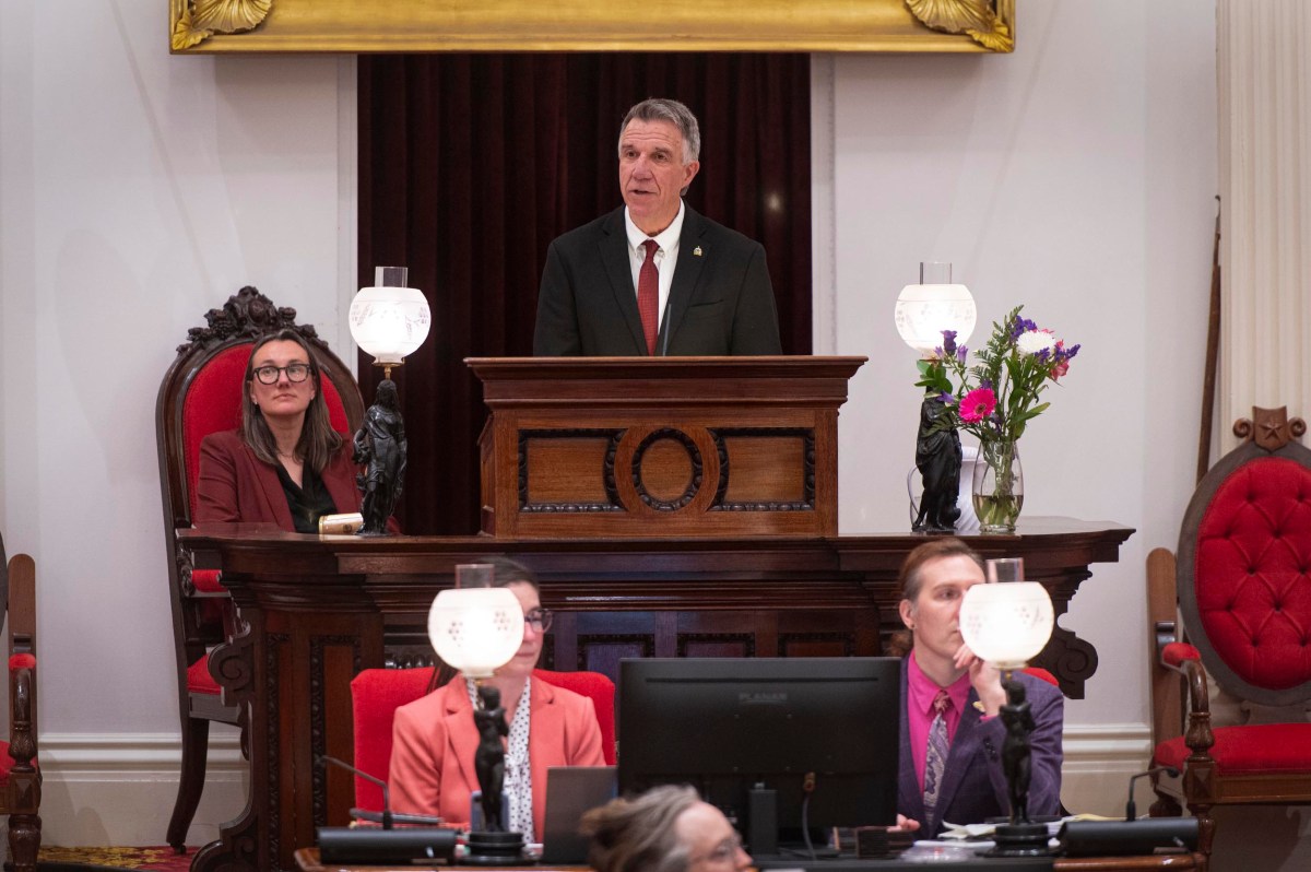 A man in a suit speaks at a podium in a formal chamber, with three seated individuals nearby and decorative lamps on the desks.