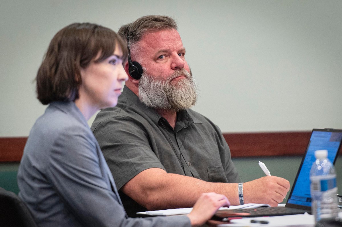 A man with headphones and a woman sit at a table with a laptop, documents, and water bottle, focused on something off camera in a meeting or courtroom setting.