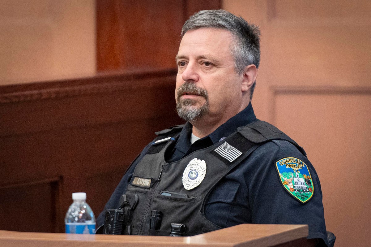 A police officer wearing a uniform and vest sits in a courtroom near a wooden surface and a bottle of water.