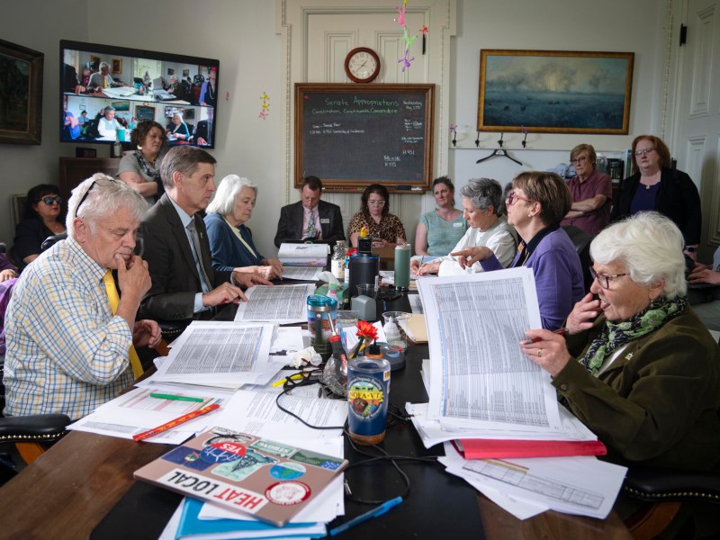 A group of people review documents around a conference table, with more participants joining via video call projected on a screen. Papers, laptops, and water bottles are scattered on the table.