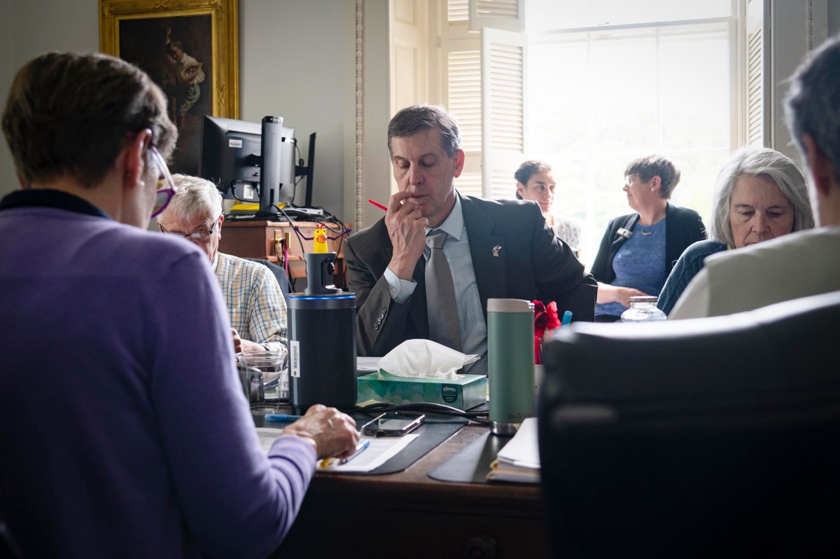 A group of people sitting around a table in a well-lit office, with papers, cups, and a computer visible; one man in a suit is holding a pencil to his lips in thought.