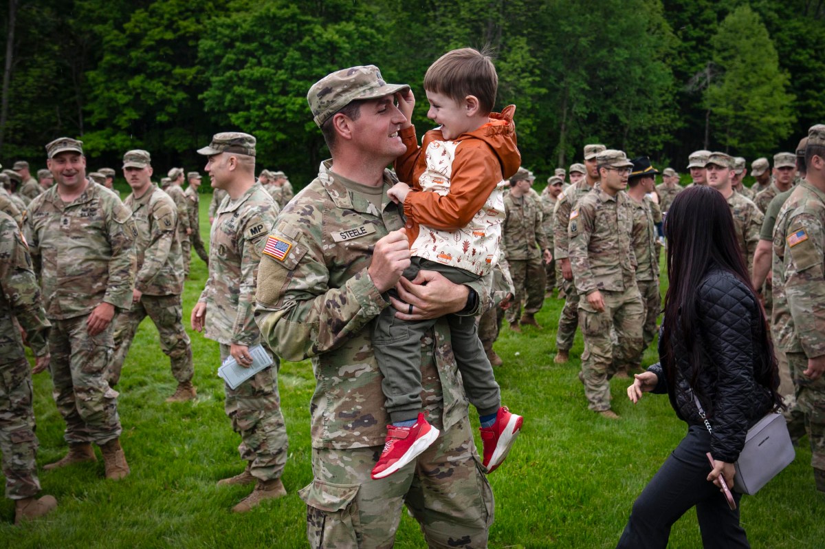 A soldier in uniform holds a young boy and smiles, surrounded by other soldiers and a few civilians on a grassy field.