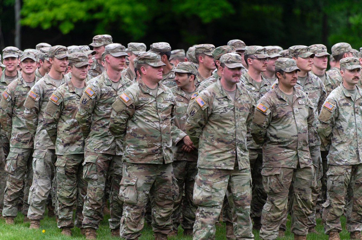 A group of U.S. Army soldiers in camouflage uniforms stand in formation outdoors on grass, with trees in the background.