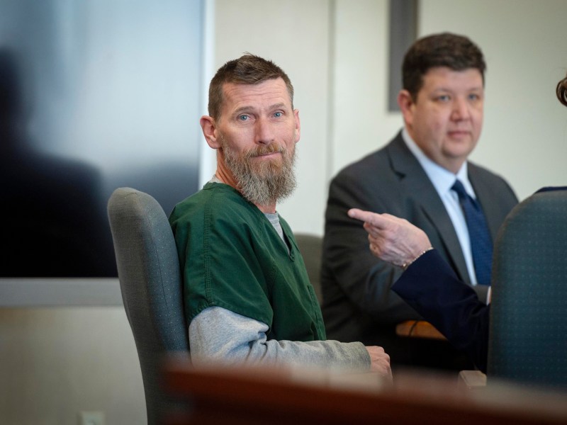 A man in a green prison uniform sits at a table in a courtroom, with two people in suits seated beside him.