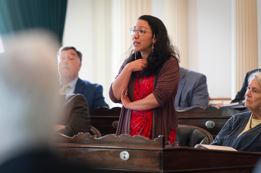 A woman in a red dress stands and speaks in a formal meeting room while others sit and listen around her.