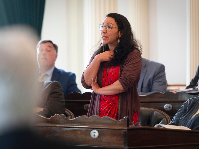 A woman in a red dress stands and speaks in a formal meeting room while others sit and listen around her.