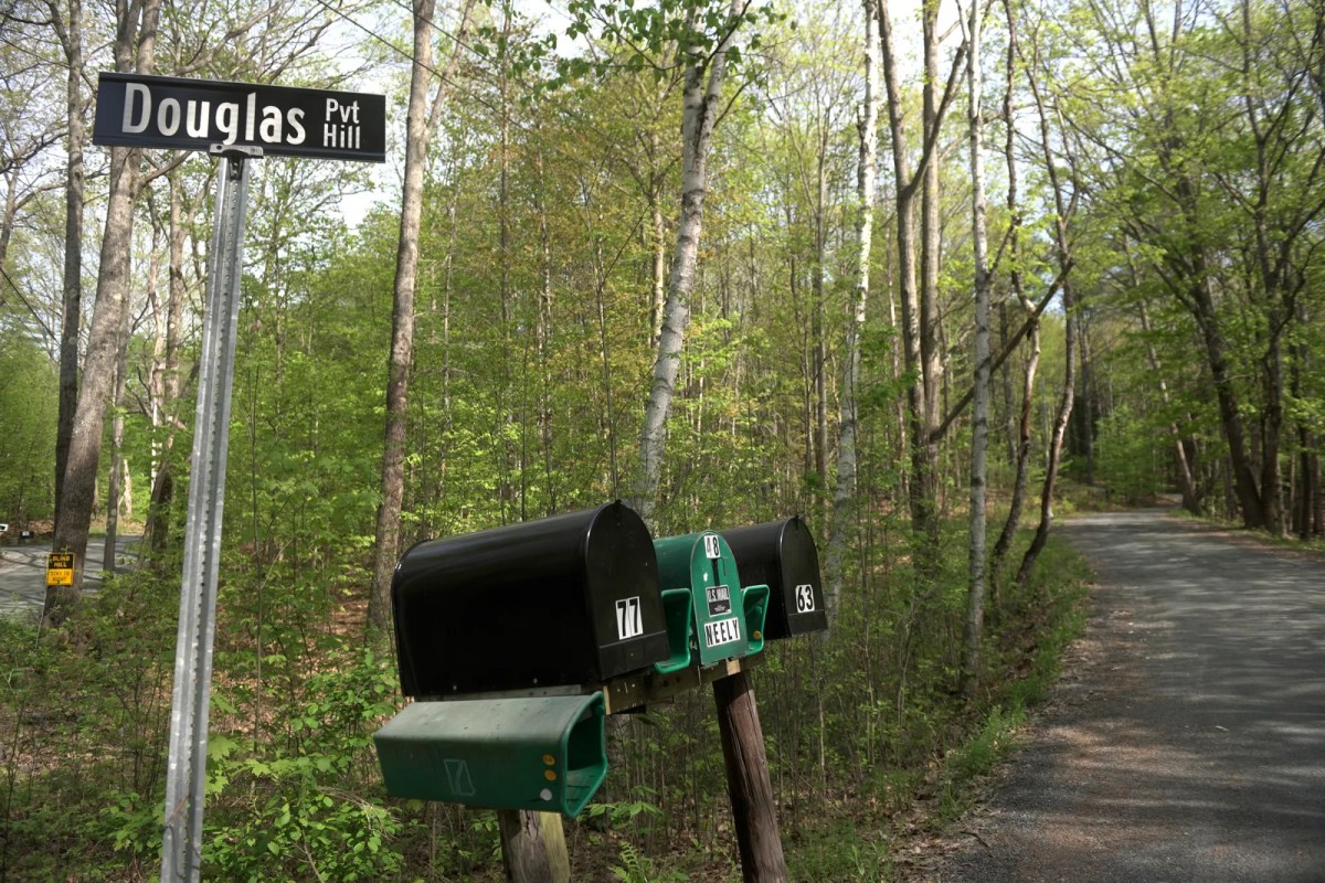A sign reading "Douglas Pvt Hill" stands next to mailboxes and a narrow paved road surrounded by green trees in a wooded area.