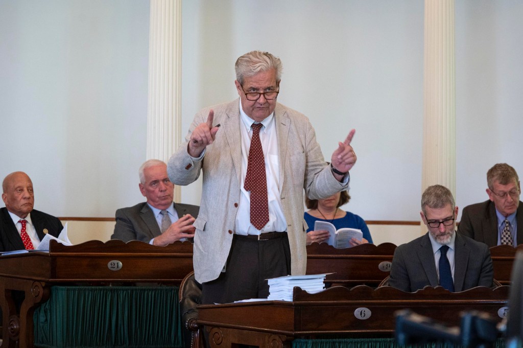 A man in a light blazer and red tie stands and gestures while speaking in a formal meeting room, with several seated people listening or reading documents.