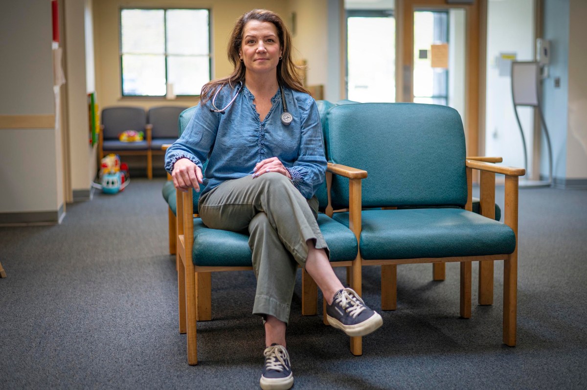 A woman with a stethoscope sits on a chair in a waiting area, looking at the camera. There are toys and chairs in the background.